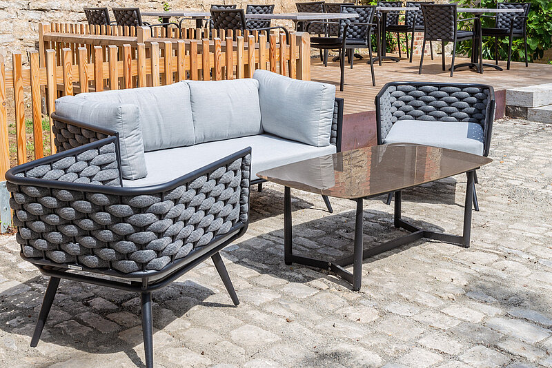 Outdoor-Lounge mit grauem, geflochtenem Sofa und Sesseln um einen Glastisch auf gepflastertem Boden